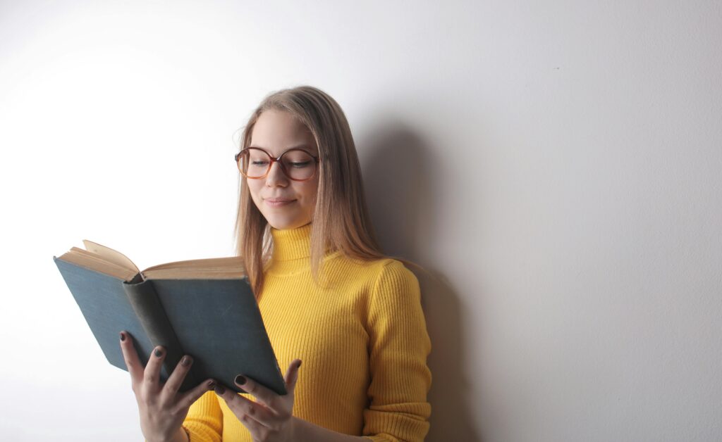 A young woman in a yellow sweater reading a book while leaning against a white wall.